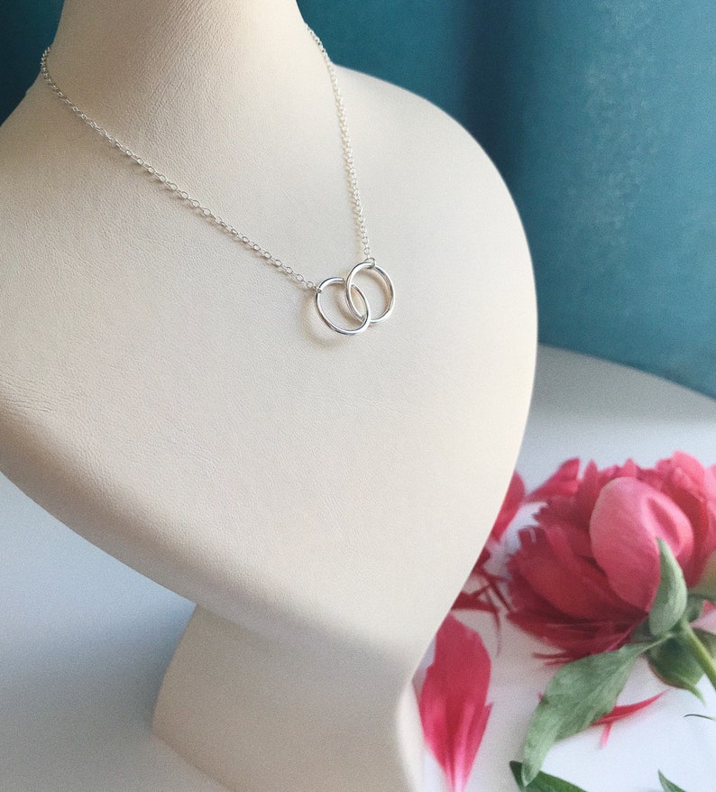 May include: A silver necklace with two interlocking circles as a pendant. The necklace is displayed on a white mannequin bust.