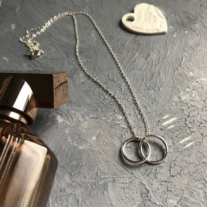 May include: A silver necklace with a double ring pendant. The necklace is on a gray textured surface.