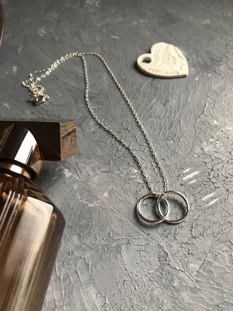 May include: A silver necklace with a double ring pendant. The necklace is on a gray textured surface.