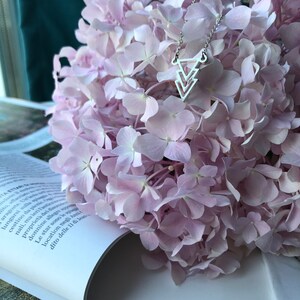 May include: A silver necklace with a geometric pendant in the shape of three overlapping triangles. The necklace is resting on a bouquet of pale pink hydrangeas.