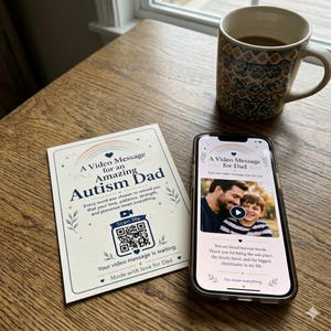 May include: A rectangular card and a smartphone displaying a video message for an amazing autism dad. The card features a rainbow, a QR code, and text. The smartphone screen shows a photo of a man and a child with a play button icon.