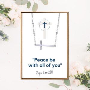 May include: A silver-colored cross necklace displayed on a white card inside a brown box. The card features a cross graphic and the text "Peace be with all of you" by Pope Leo XIV. Floral accents surround the box.
