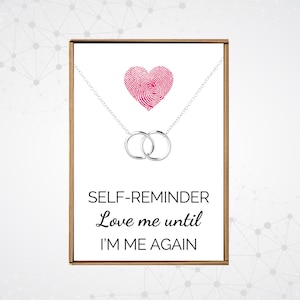 May include: A silver necklace with two interlocking rings and a pink heart charm. The necklace comes in a box with the text "SELF-REMINDER Love me until I'M ME AGAIN".