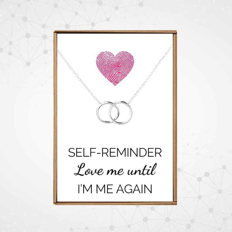 May include: A silver necklace with two interlocking rings and a pink heart charm. The necklace comes in a box with the text "SELF-REMINDER Love me until I'M ME AGAIN".