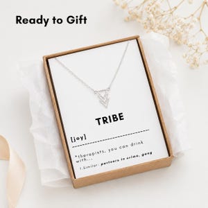 May include: A silver-toned necklace with a geometric pendant featuring three stacked triangles, presented in a brown cardboard box on a white card. The card displays the text TRIBE, [joy], and the definition: *therapists, you can drink with... 1. Similar: partners in crime, gang.