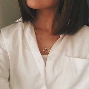 May include: A woman wearing a white button-down shirt with a delicate gold necklace featuring two interlocking circles.