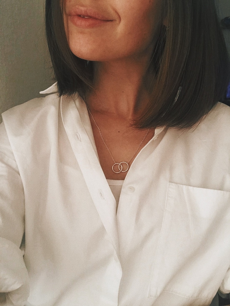May include: A woman wearing a white button-down shirt with a delicate gold necklace featuring two interlocking circles.