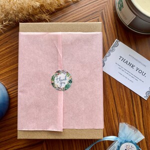 May include: A brown cardboard box with a pink tissue paper liner. The box has a sticker that says "thank you" with a floral design. A small blue bag with a white drawstring and a sticker that says "surprise!" is in the foreground.