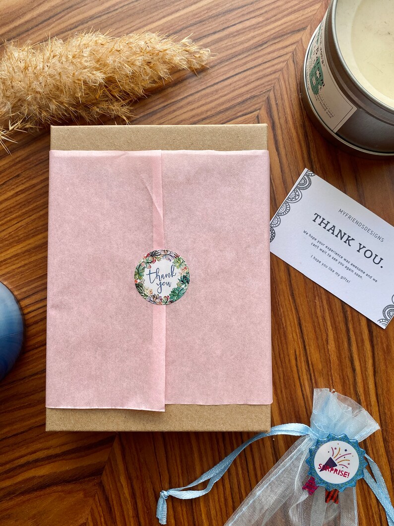 May include: A brown cardboard box with a pink tissue paper liner. The box has a sticker that says "thank you" with a floral design. A small blue bag with a white drawstring and a sticker that says "surprise!" is in the foreground.