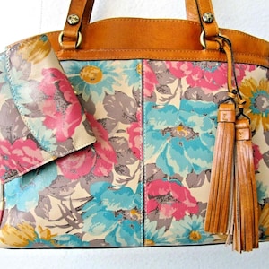 May include: Floral print handbag and matching wallet. The bag features a cream background with pink, blue, and gray flowers. It has brown leather trim, handles, and tassel accents. The wallet has a similar floral pattern.