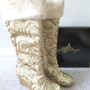 May include: A pair of knee-high boots with a wedge heel. The boots are covered in gold sequins with a swirling pattern and topped with a fluffy, cream-colored faux fur trim. A black box with gold lettering is in the background.