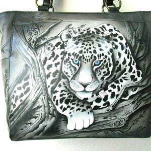 May include: A gray tote bag featuring a detailed illustration of a leopard with blue eyes, set against a backdrop of trees and foliage. The leopard's spots are black and white, and the bag has black handles.