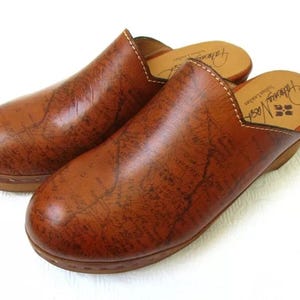 May include: A pair of brown leather clogs with a map-like pattern. The clogs have a rounded toe and a wooden sole. The interior of the clogs has a brand name printed on the insole. The clogs are made of Italian leather.