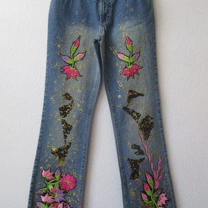 May include: Blue denim jeans with embroidered floral designs in pink, green, and gold. The jeans have a bootcut leg style.