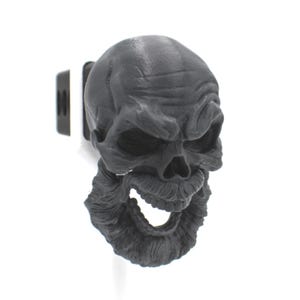 May include: A matte black hitch cover featuring a detailed, three-dimensional human skull with a prominent mustache and a full, textured beard. The skull has deep-set eye sockets, a defined nose cavity, and an open mouth, attached to a square mount.