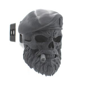 May include: A matte black hitch cover featuring a sculpted skull wearing a military-style beret with a small emblem on the side. The skull has a detailed beard and a cigar held between its teeth, attached to a rectangular metal mounting bracket.