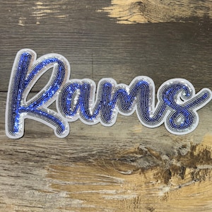 May include: A sequined patch with the word "Rams" in a cursive font. The letters are outlined in silver sequins and filled with blue sequins. The patch is on a wooden background.