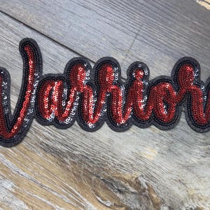 May include: A sequined patch in the shape of the word "Warriors" in a cursive font. The letters are filled with red sequins and outlined with black sequins and a black border. The patch is on a wooden surface.