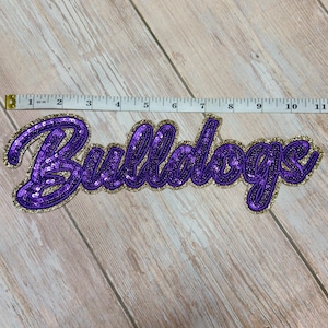 Puede incluir: Un parche de lentejuelas moradas con la palabra Bulldogs en letra cursiva y un borde de lentejuelas doradas. Una cinta métrica blanca está colocada sobre el parche, mostrando una longitud total de aproximadamente 29,2 cm para el diseño.