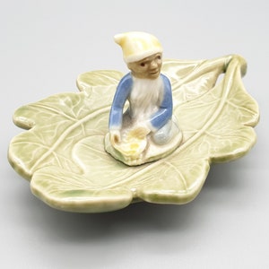 May include: A small ceramic figurine of a gnome sitting on a green leaf-shaped dish. The gnome is wearing a blue and white outfit and a yellow hat.