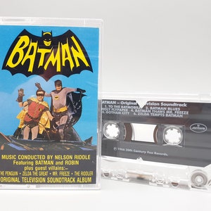 Könnte beinhalten: Eine Vintage-Kassette mit dem Soundtrack der Batman-Fernsehserie. Das Cover zeigt eine schwarz-gelbe Illustration von Batman und Robin, die auf dem Batmobil stehen. Der Text auf dem Cover lautet "BATMAN" und "Original Television Soundtrack Album".