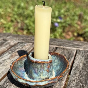May include: A ceramic candle holder with a teal and brown glaze holds a cream-colored candle. The candle holder has a small dish and a central cup to hold the candle. The candle is unlit.