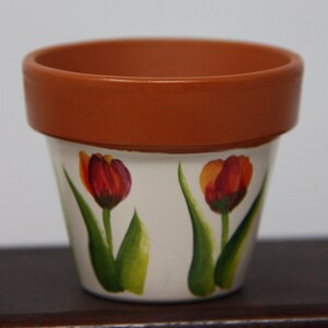 Puede incluir: Una maceta de terracota con base blanca pintada con dos tulipes rojas y naranjas con hojas verdes.