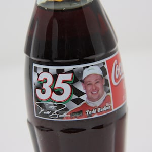 May include: A Coca-Cola bottle with a label featuring NASCAR driver Todd Bodine. The label has a checkered flag background with the number 35 and Bodine's signature.