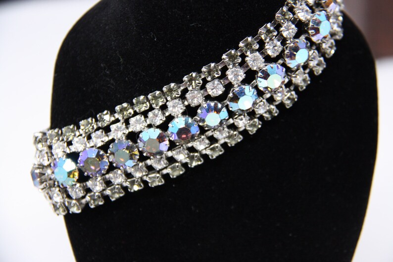 May include: A silver and crystal rhinestone bracelet with iridescent stones. The bracelet is on a black velvet display stand.