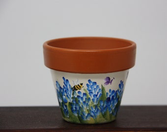 May include: A terracotta flower pot with a white background and a painted design of blue flowers, green leaves, a yellow and black bee, and purple butterflies.