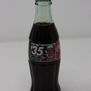 May include: A glass bottle of Coca-Cola with a red cap and a label featuring a race car driver with the number 35 and the name "Todd Bodine".