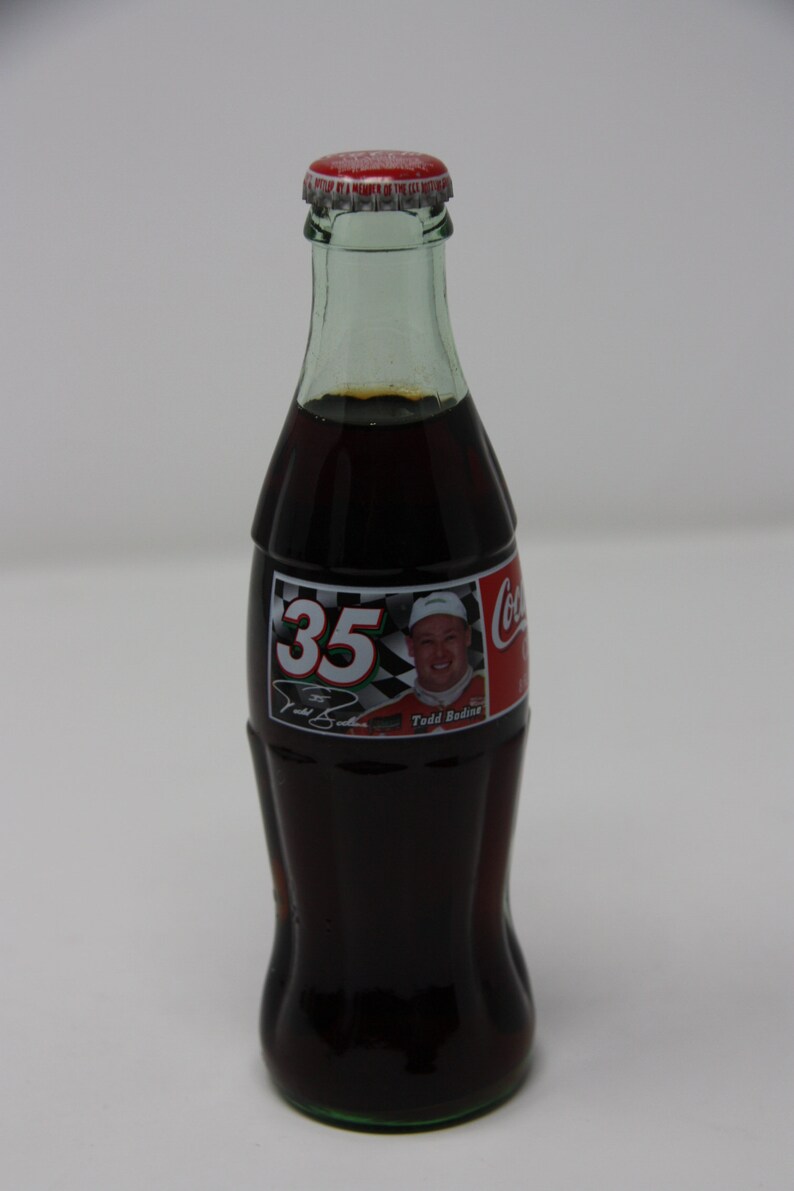 May include: A glass bottle of Coca-Cola with a red cap and a label featuring a race car driver with the number 35 and the name "Todd Bodine".