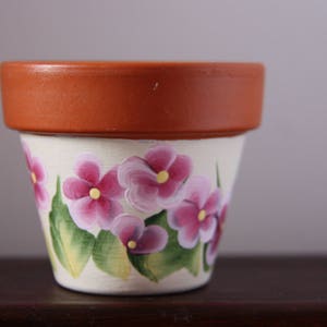 Puede incluir: Una maceta de terracota con base blanca pintada con flores rosas y hojas verdes.