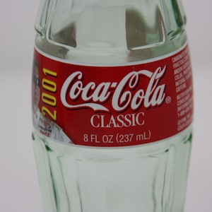 May include: A clear glass Coca-Cola bottle with a red label featuring the Coca-Cola logo and the text "CLASSIC". The label also includes the year "2001" and the volume "8 FL OZ (237 mL)".