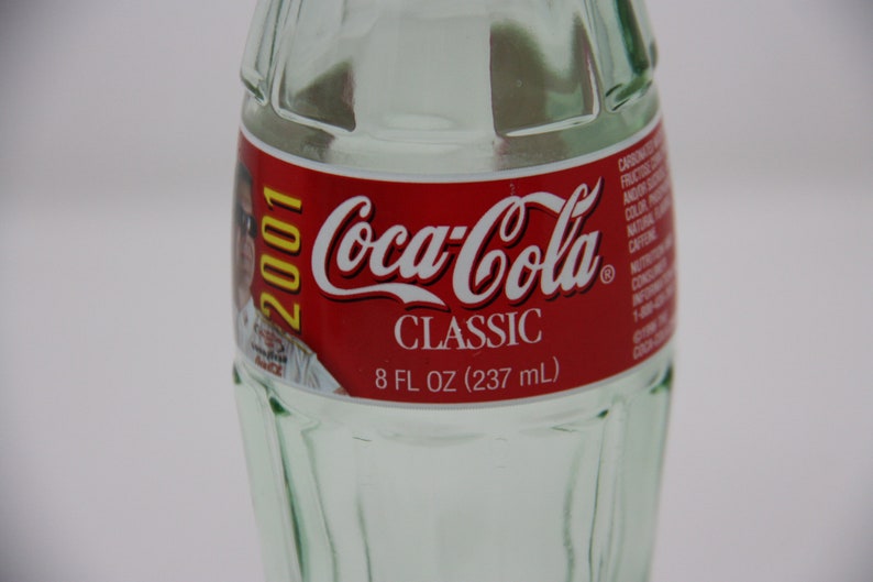 May include: A clear glass Coca-Cola bottle with a red label featuring the Coca-Cola logo and the text "CLASSIC". The label also includes the year "2001" and the volume "8 FL OZ (237 mL)".