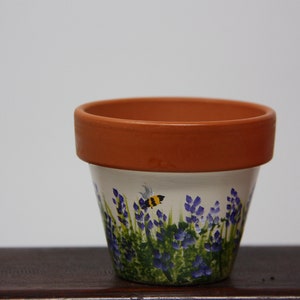 Op de afbeelding: Een terracotta bloempot met een witte achtergrond en een geschilderd ontwerp van paarse bloemen en een gele en zwarte bij.