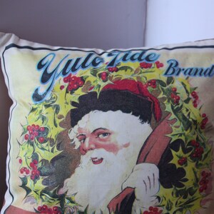 May include: A white pillow with a vintage illustration of Santa Claus wearing a red hat and holding a bag. The illustration is surrounded by a wreath of holly with red berries. The text "Yule Tide Brand" is printed in blue and black lettering.
