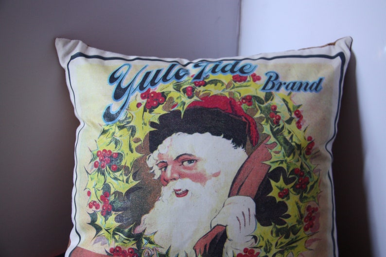 May include: A white pillow with a vintage illustration of Santa Claus wearing a red hat and holding a bag. The illustration is surrounded by a wreath of holly with red berries. The text "Yule Tide Brand" is printed in blue and black lettering.
