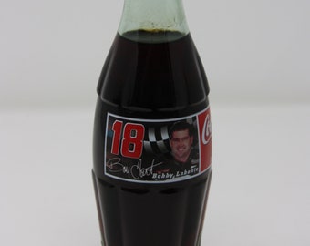 May include: A glass bottle of Coca-Cola with a red and white label featuring a photo of race car driver Bobby Labonte. The label also includes the number 18 and the text "Boy Scout".