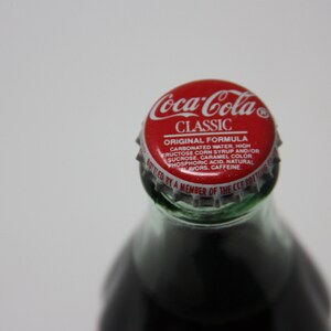 May include: A red and white Coca-Cola Classic bottle cap with the text "Coca-Cola Classic Original Formula Carbonated Water High Fructose Corn Syrup and/or Sucrose, Caramel Color Phosphoric Acid, Natural Flavors, Caffeine Bottled by a Member of the CCE Bottling Group" printed on it.