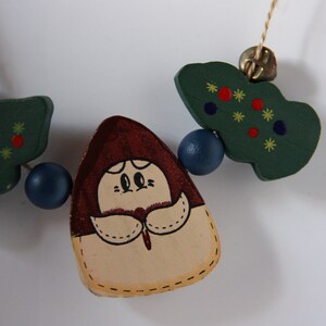 May include: A wooden garland featuring a brown and cream figure with a red hat, two green trees with red and yellow stars, and blue beads.