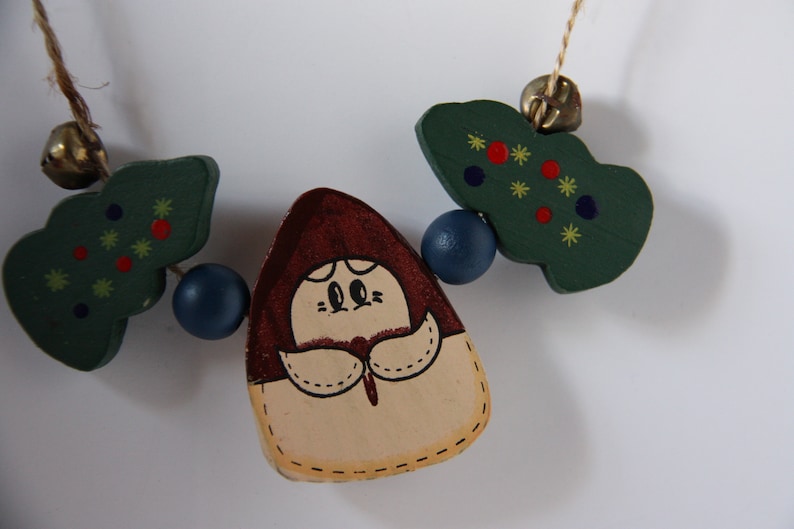 May include: A wooden garland featuring a brown and cream figure with a red hat, two green trees with red and yellow stars, and blue beads.