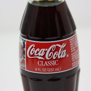 May include: A glass bottle of Coca-Cola Classic, 8 fl oz (237 ml). The bottle has a red label with the Coca-Cola logo and the words "Classic" and "8 fl oz (237 ml)".