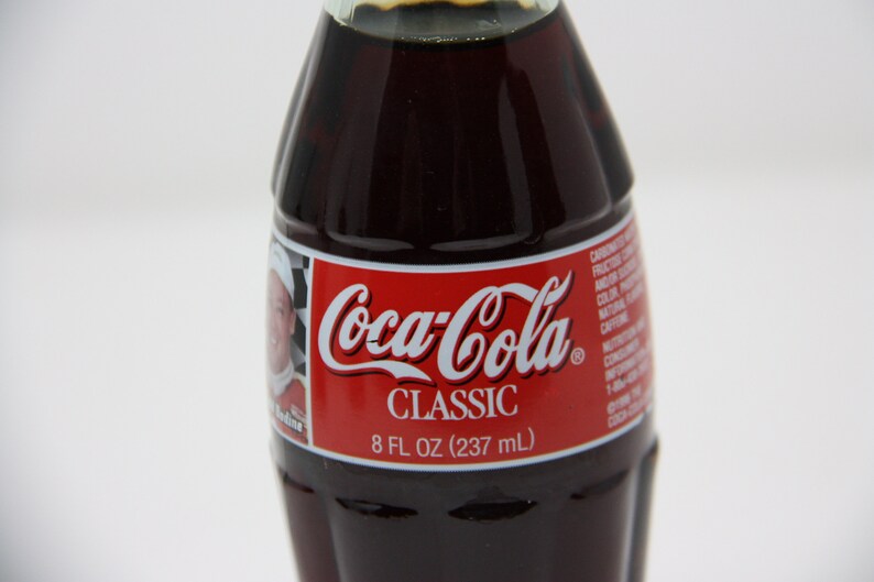 May include: A glass bottle of Coca-Cola Classic, 8 fl oz (237 ml). The bottle has a red label with the Coca-Cola logo and the words "Classic" and "8 fl oz (237 ml)".
