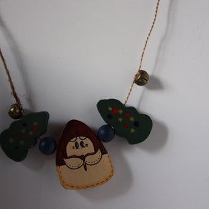 May include: A wooden Christmas garland featuring a girl figure in the centre, surrounded by green trees with red and yellow accents, and blue and gold bells.