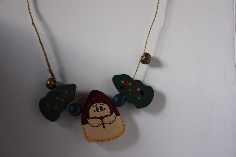May include: A wooden Christmas garland featuring a girl figure in the centre, surrounded by green trees with red and yellow accents, and blue and gold bells.