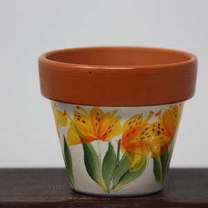 May include: A terracotta flower pot with a white base painted with orange and yellow lilies.