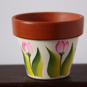 Op de afbeelding: Een terracotta bloempot met een witte basis beschilderd met roze tulpen en groene bladeren.