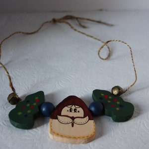 May include: A wooden Christmas decoration featuring a painted image of a woman in a red dress and white apron. The decoration is flanked by two green trees with red and yellow decorations. The ornament is strung on a brown twine.