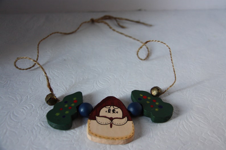 May include: A wooden Christmas decoration featuring a painted image of a woman in a red dress and white apron. The decoration is flanked by two green trees with red and yellow decorations. The ornament is strung on a brown twine.